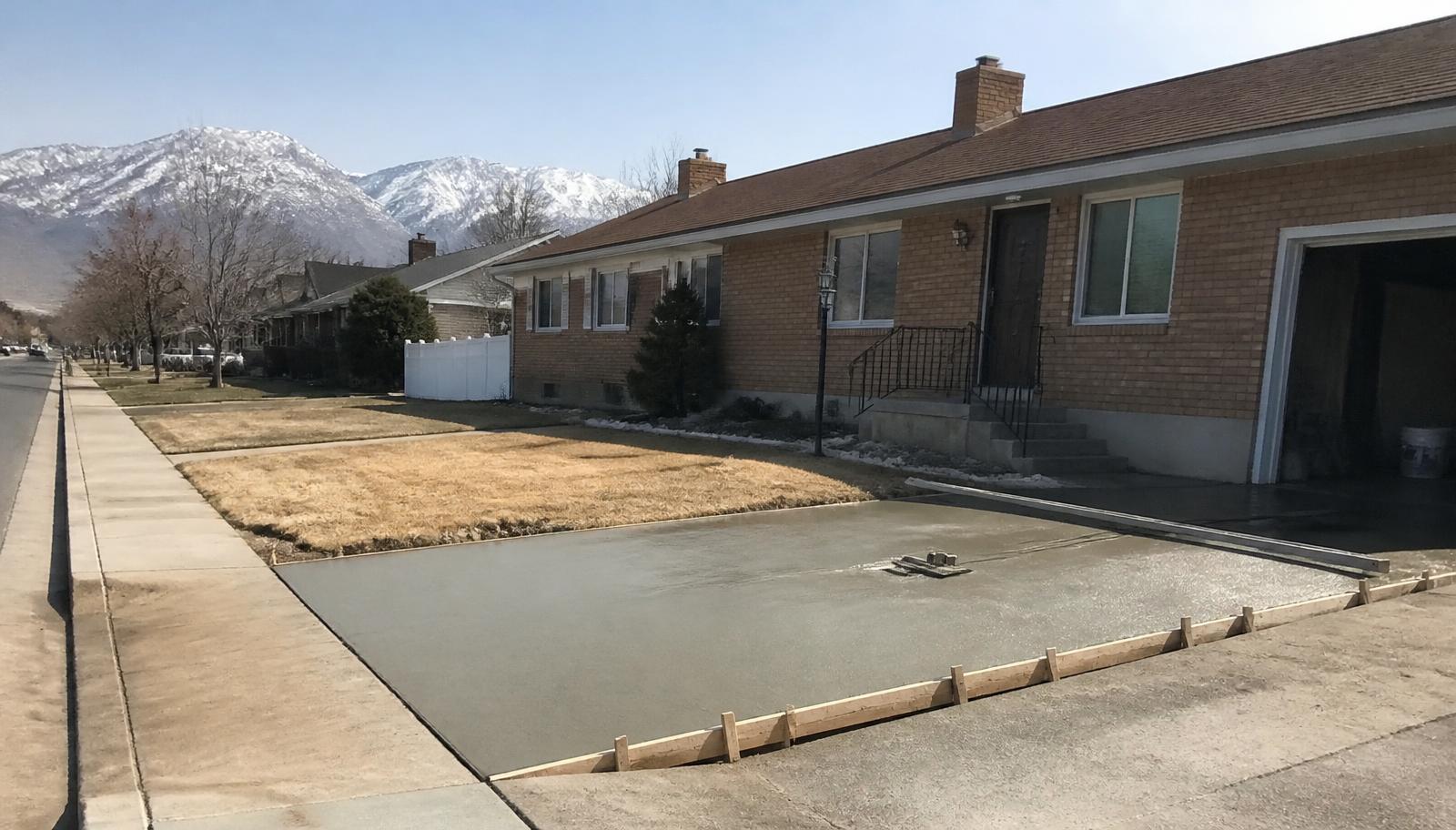 Freshly poured concrete driveway apron curing beside a brick rambler with snowy mountains in the distance