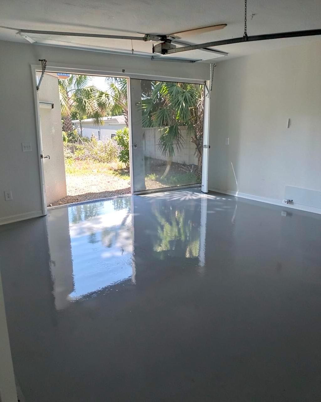 Glossy gray epoxy-coated garage floor in an empty residential garage with the door open to palm shadows