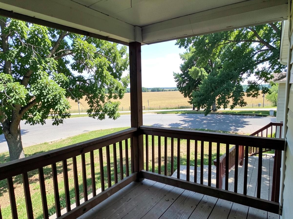 Simple covered front porch with wooden railing looking out over a quiet street toward flat open country