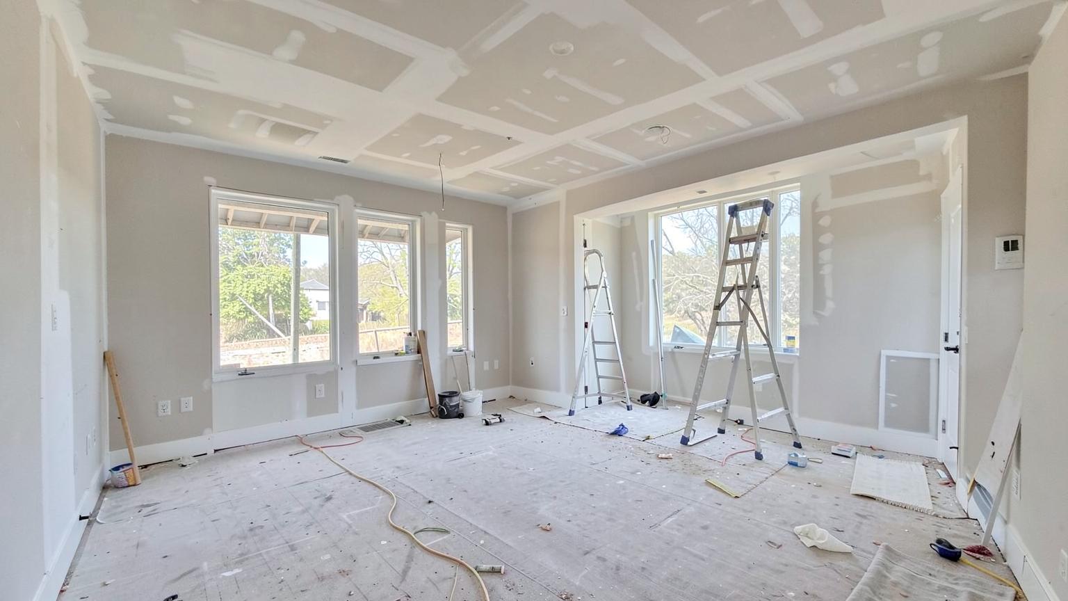 Interior of a new home during finishing work with fresh drywall, bare floors, ladder and large bright windows