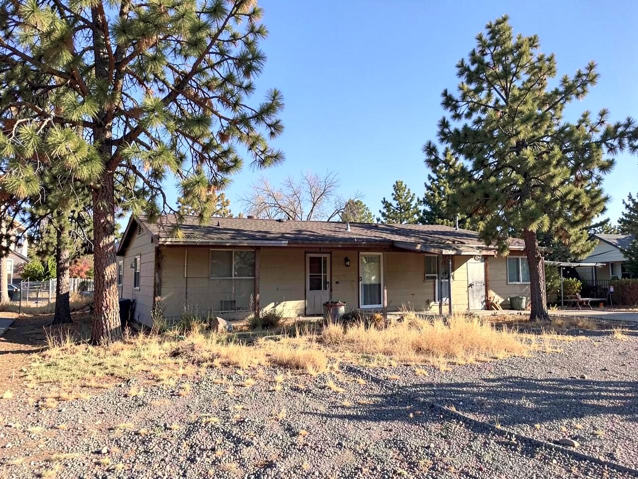 Older single-story home with a low roofline, mature pines, and a dry gravel yard in high-desert light