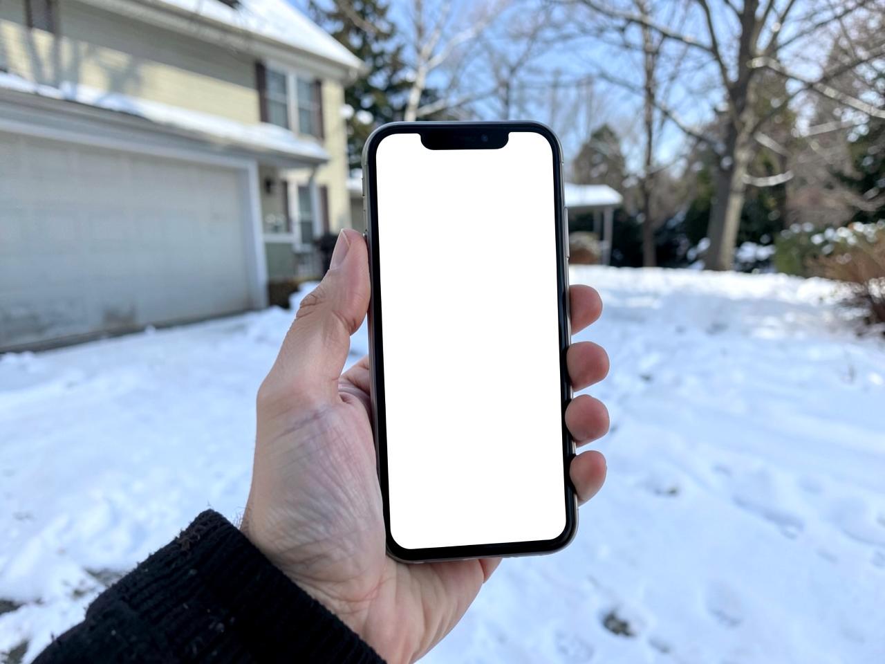 Hand holding a phone with a blank screen while standing on a snowy driveway in front of a house