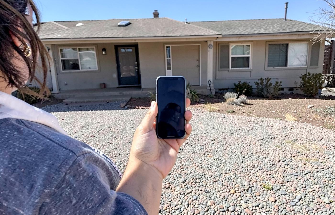 Person holding a smartphone in a gravel desert front yard in front of a modest single-story home