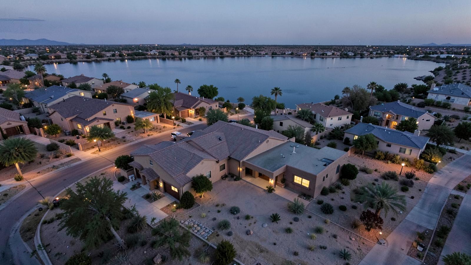 Aerial dusk view of a desert lakeside neighbourhood of low stucco homes with warm street lights and calm water beyond