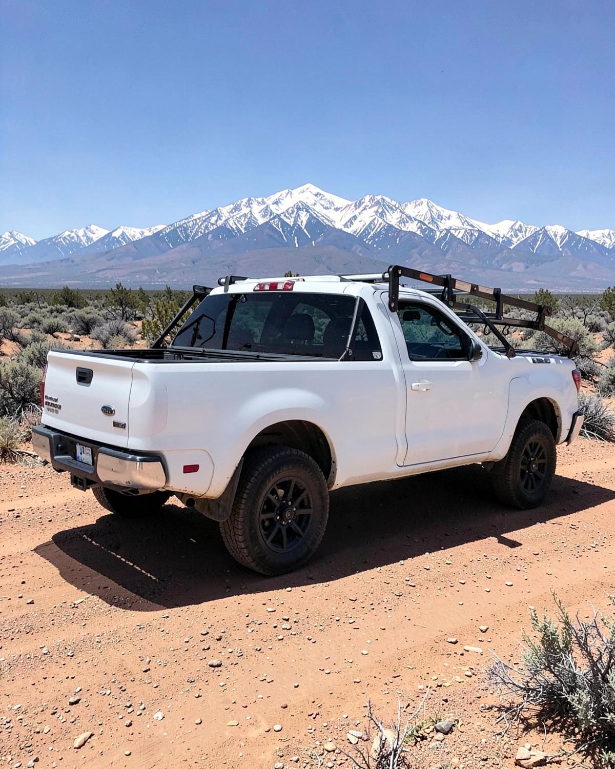 Plain white contractor pickup truck with a ladder rack parked on a dirt track in open sagebrush country below snow-capped peaks