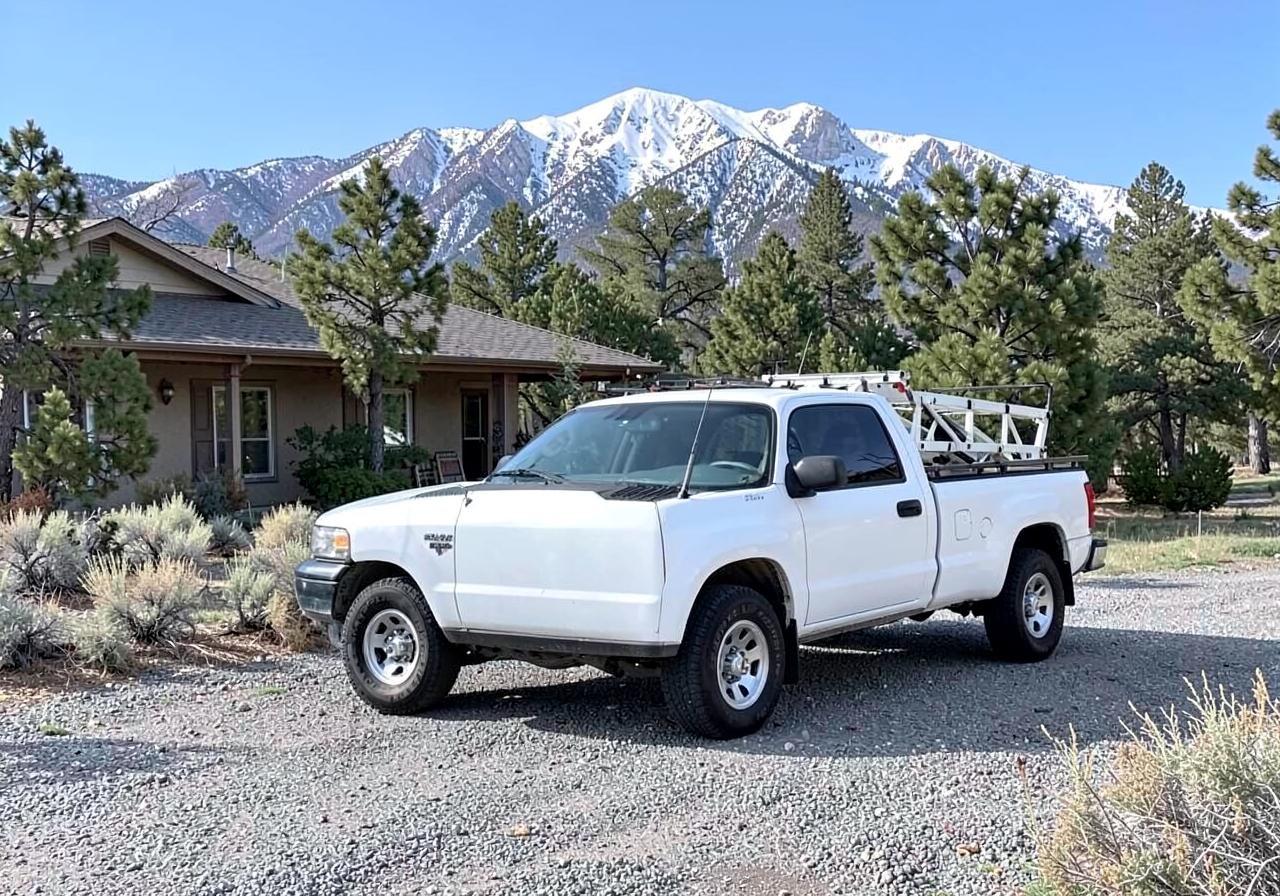 White unmarked contractor pickup with a ladder rack parked on a gravel driveway of a modest home with snowy mountains behind