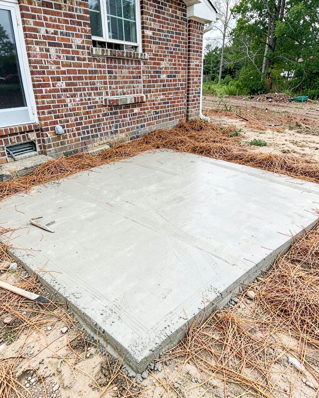 Freshly finished concrete patio slab behind a brick home surrounded by pine straw and sandy soil