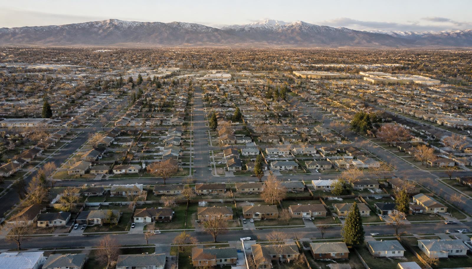 Aerial view of a dense flat grid of modest suburban rooftops in a broad valley with a long mountain ridge on the horizon